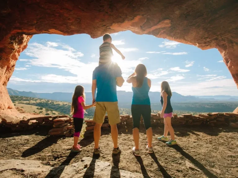 A family of five stands in a rocky cave opening, overlooking a scenic landscape with hills and a bright blue sky. The father holds a young child on his shoulders, and sunlight filters through the archway. Travel Destination Ideas With 4-Year-Old