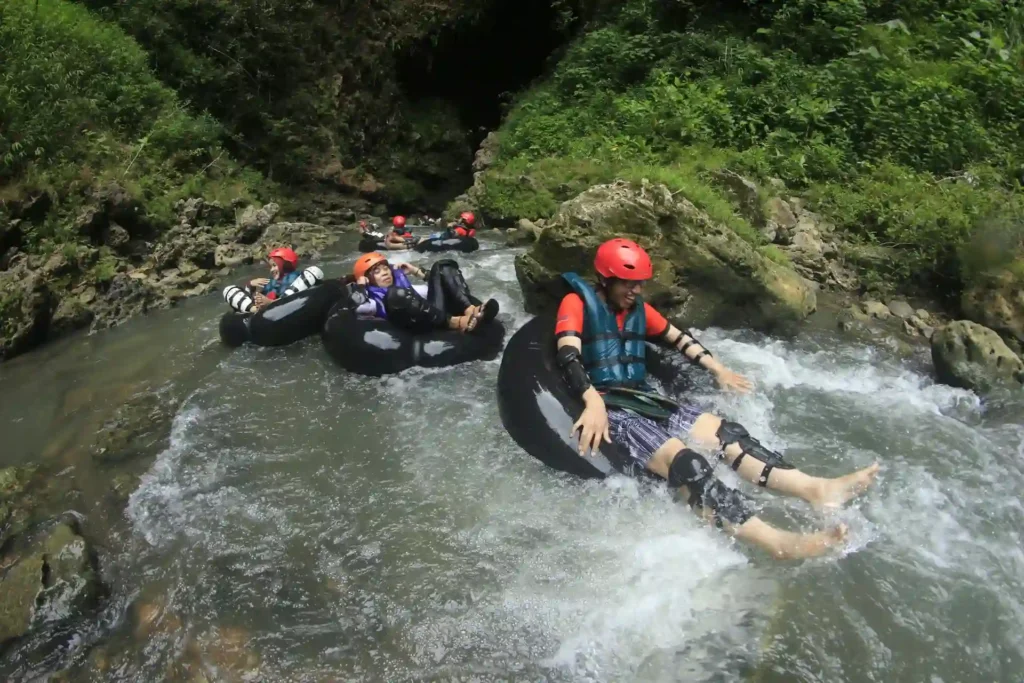 A group of people with helmets and life jackets are tubing down a river in inner tubes, enjoying the thrills of adventure travel. They navigate through rocky areas surrounded by lush greenery. The water appears to be flowing steadily, creating small splashes as they move through the river.