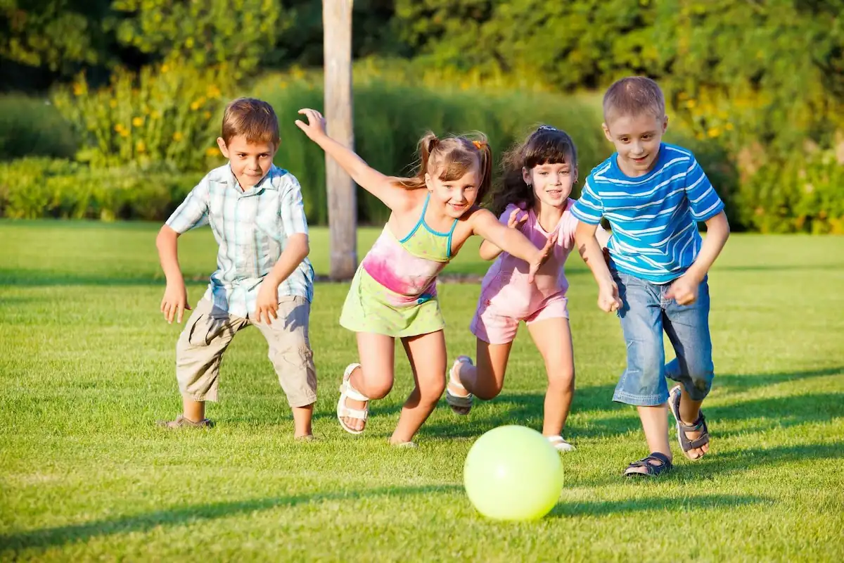 Four children are playing outside on a lush green lawn. They are all smiling and appear to be racing towards a green ball in front of them. The background features tall grass and trees, suggesting a park or garden setting. The children are dressed in summer clothes. Affordable Activities for Kids in Texas