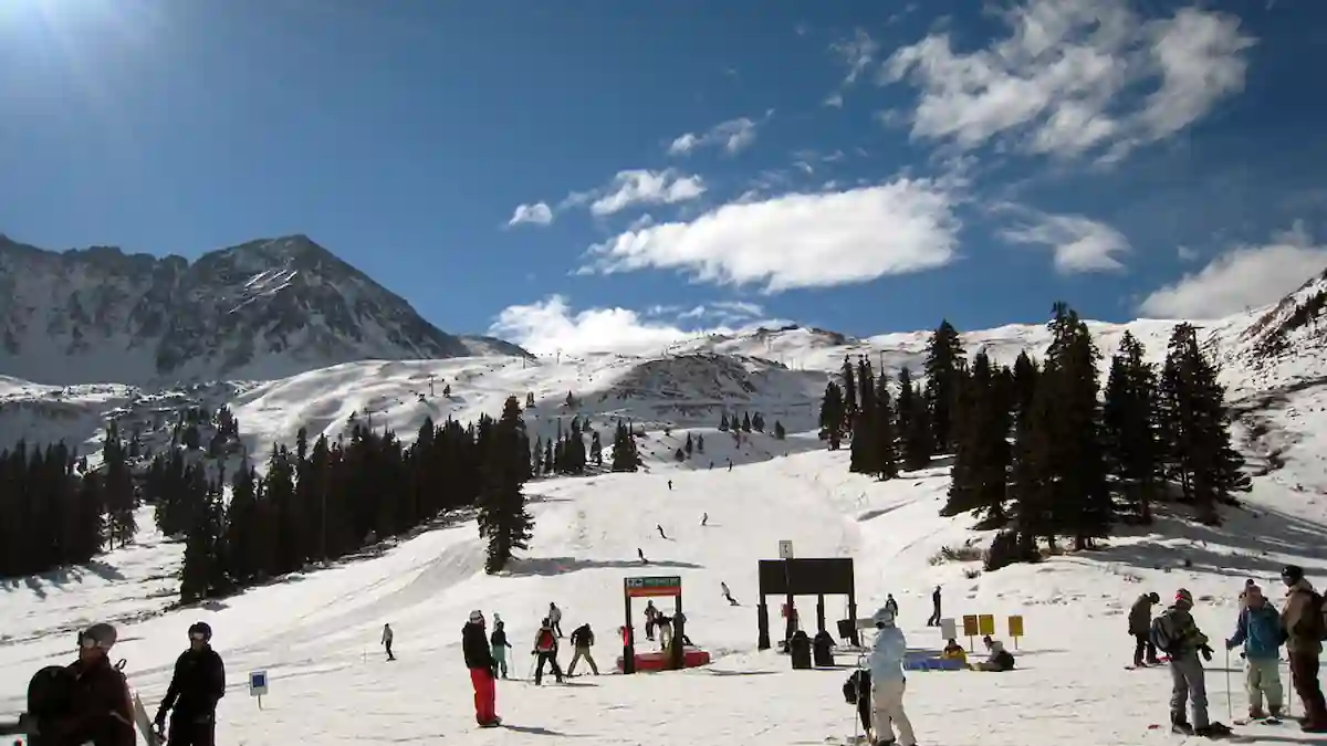 A scenic mountain landscape features a busy ski resort with snowy slopes. Skiers and snowboarders are seen enjoying the slopes, while pine trees dot the scenery. A bright, sunny sky with scattered clouds enhances the picturesque setting.
