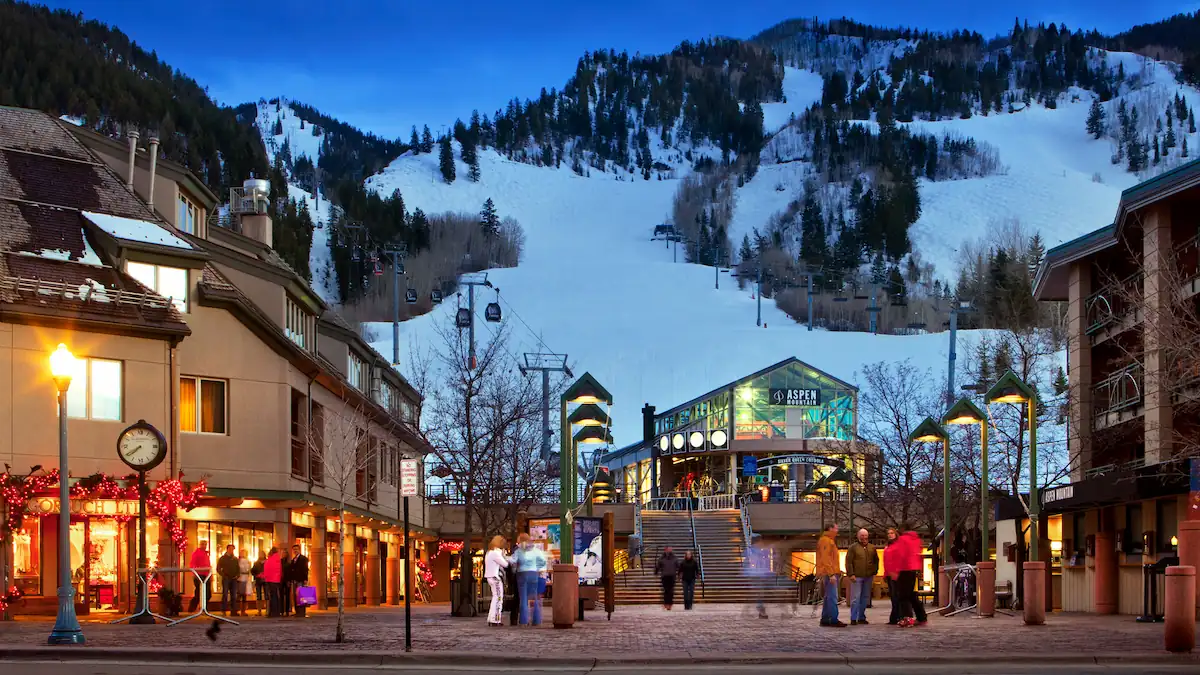 A lively winter evening in Aspen featuring the entrance to a ski resort with snow-covered mountains in the background. People are gathered near shops adorned with festive decorations, and a lit-up staircase leads up to the gondola station. Ikon Pass Destinations