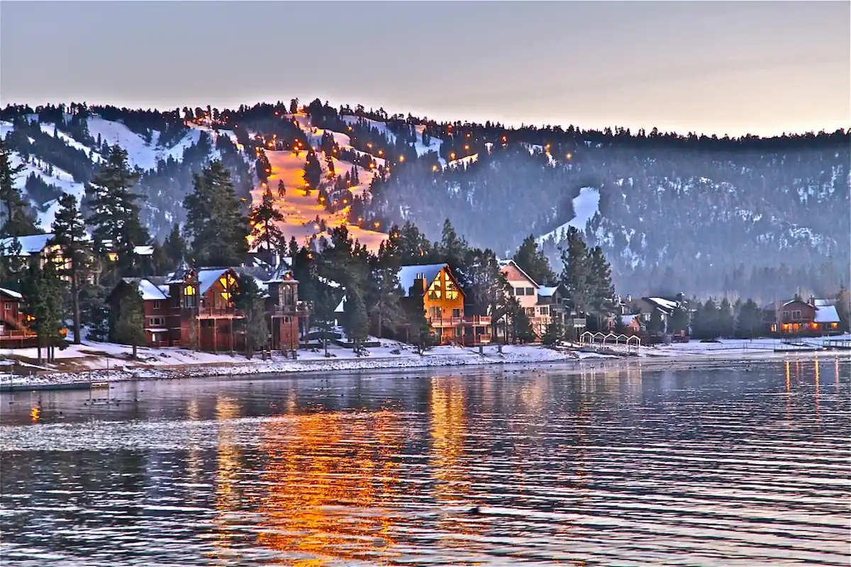 A serene lakeside village at dusk, with snow-covered houses nestled among tall pine trees. In the background, a snowy mountain with illuminated ski slopes reflects warm lights onto the calm water, creating a peaceful and picturesque winter scene.
