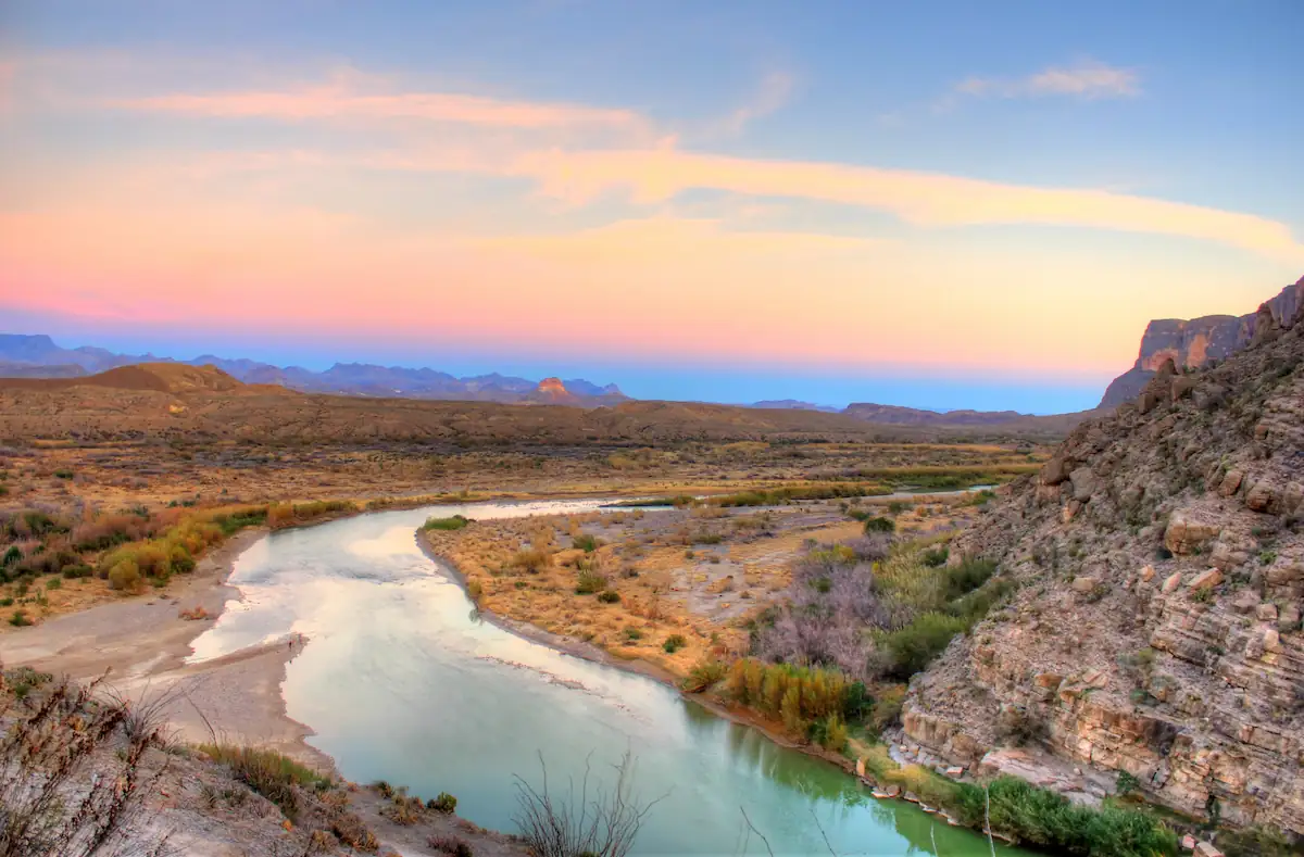 A serene landscape featuring a river winding through a rugged, arid terrain at sunset. The sky displays a gradient of colors from blue to pink, casting a warm glow over the distant hills and rocky cliffs lining the riverbanks. Sparse vegetation dots the scene.
