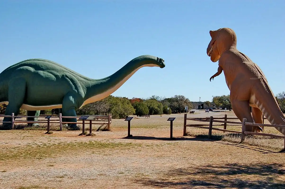 Two large dinosaur sculptures stand in a fenced outdoor area. The green one on the left is a long-necked sauropod, and the tan one on the right is a bipedal theropod. Information plaques are in front of each statue, and trees and buildings are visible in the background.
