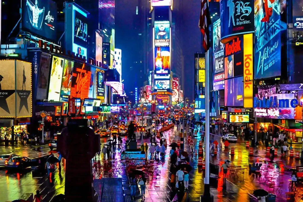 A vibrant, rainy night in Times Square, New York City, with colorful, glowing billboards illuminating the bustling crowds and wet streets. Pedestrians with umbrellas and raincoats walk by, discussing plans to enjoy a Broadway show while cars and taxis navigate the busy roads.