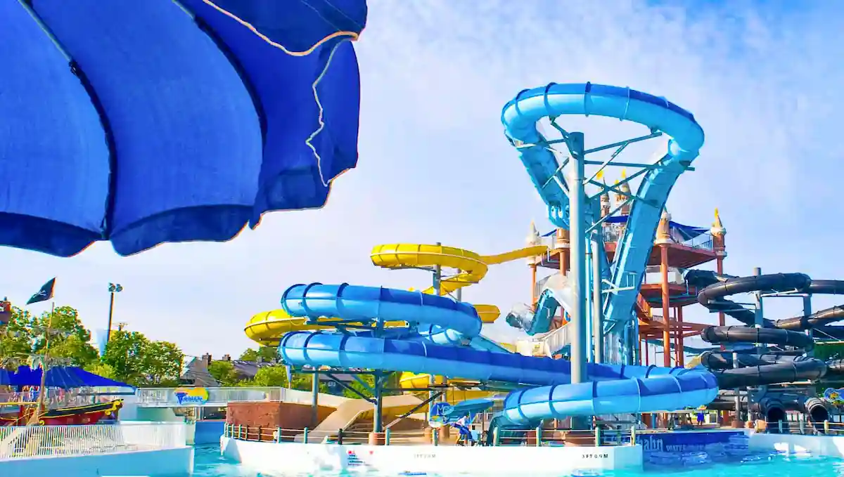 A vibrant water park featuring multiple twisting waterslides in blue, yellow, and black, with pools at the bottom. An overhead blue shade structure is partially visible. The clear, sunny sky is in the background, emphasizing a fun, summer atmosphere.
