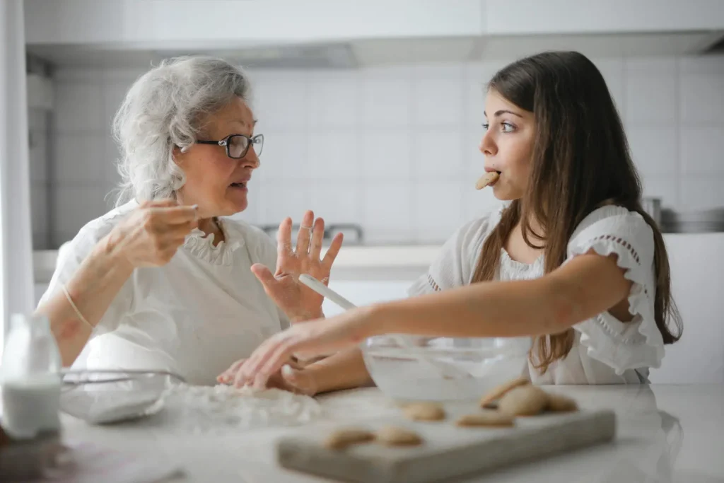 An elderly woman and a young girl are in a kitchen, presumably baking cookies together. The girl is eating a cookie while the elderly woman offers expert advice, gesturing animatedly. The table is filled with baking items like a bowl, dough, and milk. Both are wearing white tops.