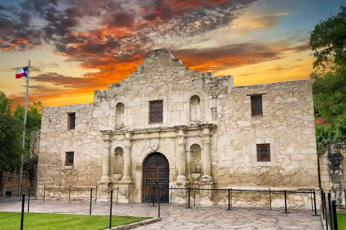 A historic stone mission stands under a vibrant sunset sky with wisps of orange, red, and yellow clouds. The mission features an arched entrance, wooden doors, and small windows. A flagpole with a colorful flag is visible on the left side of the image.
