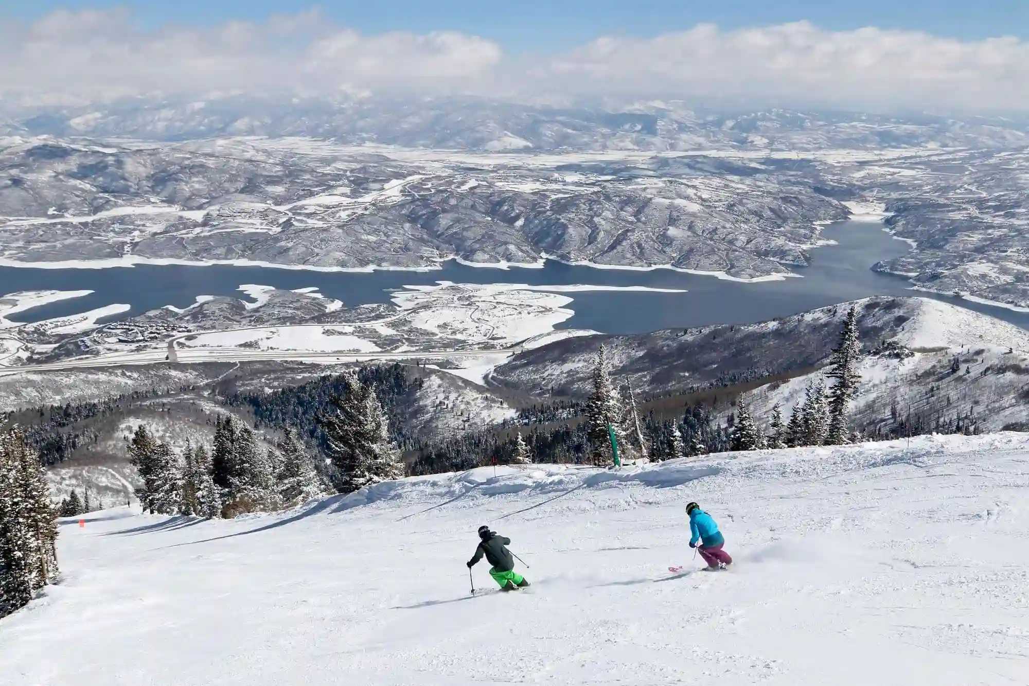 Two skiers, one in green pants and the other in blue, glide down a snowy mountain slope with trees alongside. In the background, a scenic view of Ikon Pass Destinations reveals snow-covered mountains and a lake under a cloudy sky.