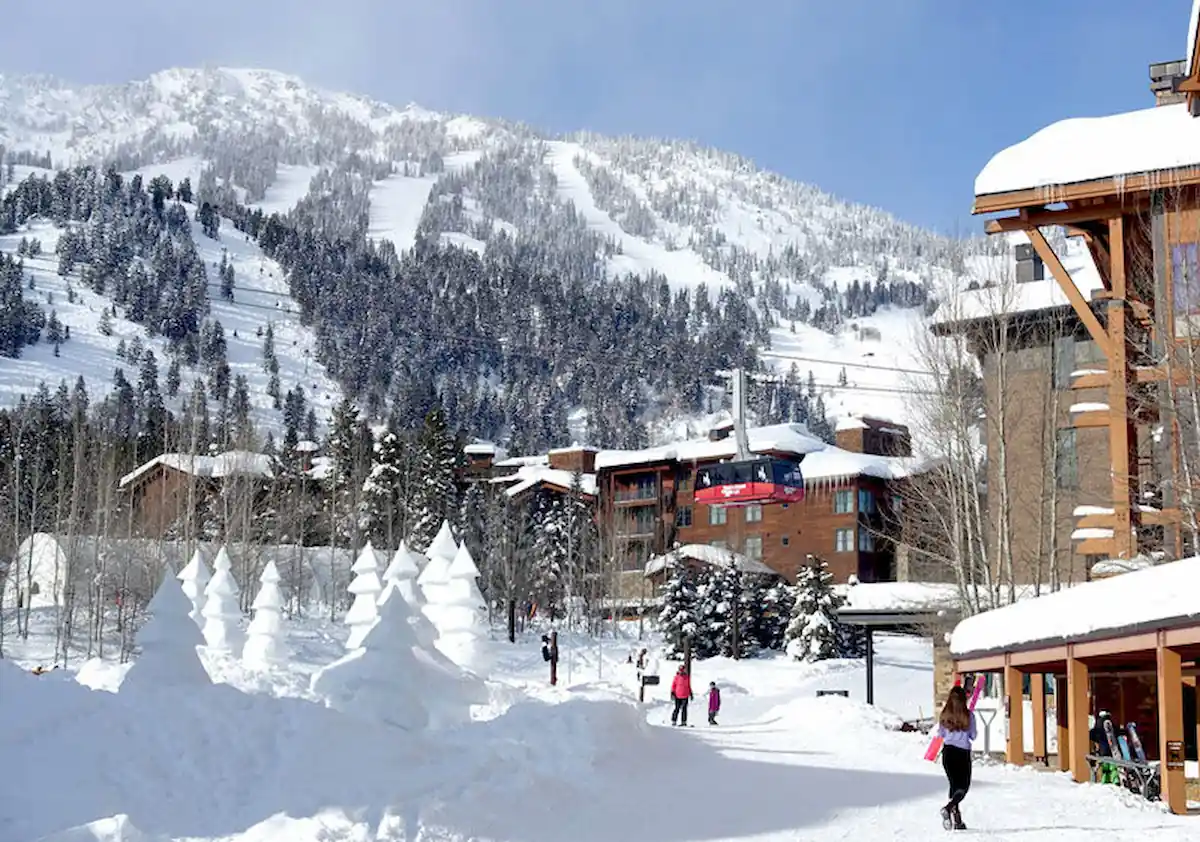 A snowy ski resort with people walking, trees and buildings covered in snow. A red cable car is ascending towards a snow-covered mountain in the background. Pine trees and festive decorations enhance the wintry scene.
