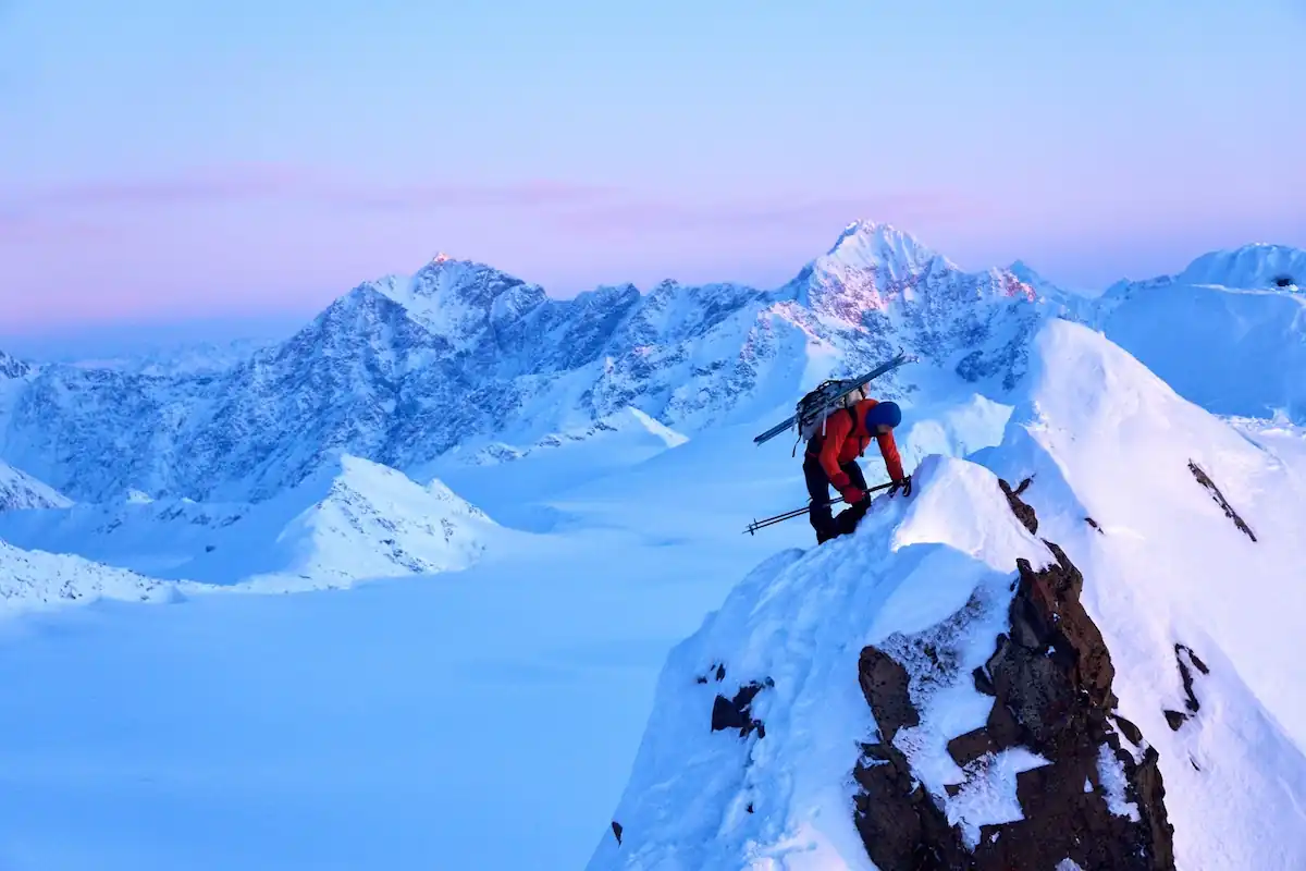 A climber in red and blue gear ascends a snowy peak carrying skiing equipment. The background features a panoramic view of snow-covered mountains under a pastel-colored sky at either dawn or dusk.
