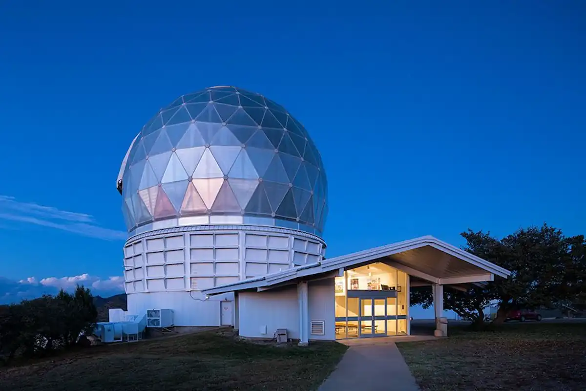 A large geodesic dome housing an observatory stands against a twilight sky. The dome is lit from within, emitting a soft glow. Next to it is a smaller, white building with a slanted roof and glass doors, also illuminated from inside. Trees and hills surround the structures.
