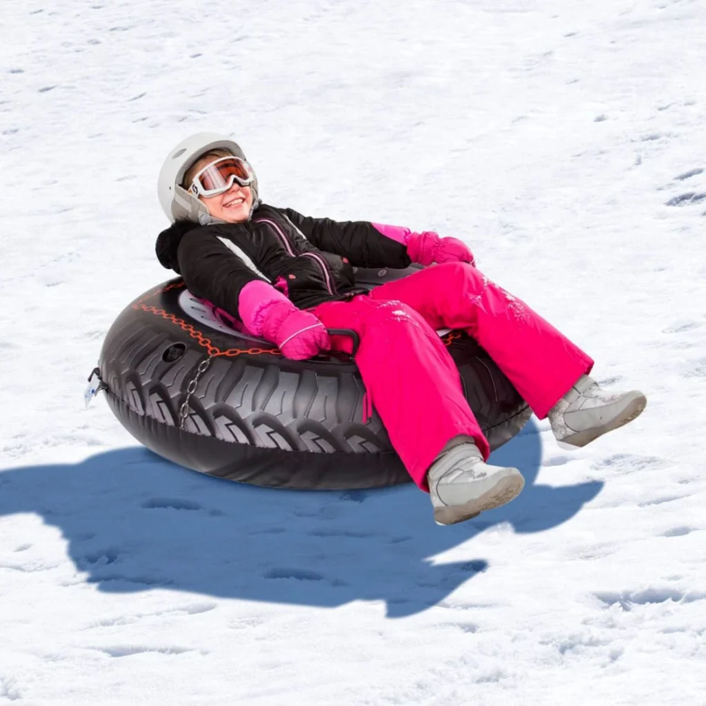 A child wearing a white helmet, goggles, a black jacket, pink pants, and white boots is sitting on a black and gray snow tube with tire tread design. The child appears to be having fun sliding down a snowy slope at Mount Peter.