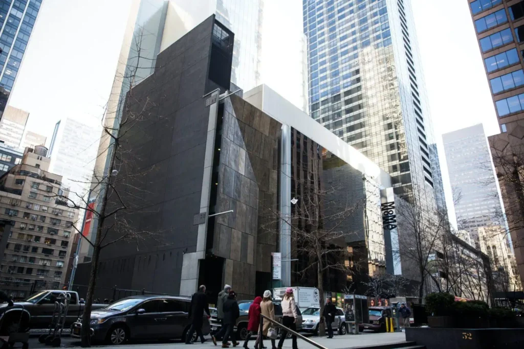 Street view of a modern building with dark and reflective façades, surrounded by other high-rise structures. People walk on the sidewalk in various directions, some crossing the street. A few cars and a white van are parked along the road in front of what is now the Museum of Modern Art.