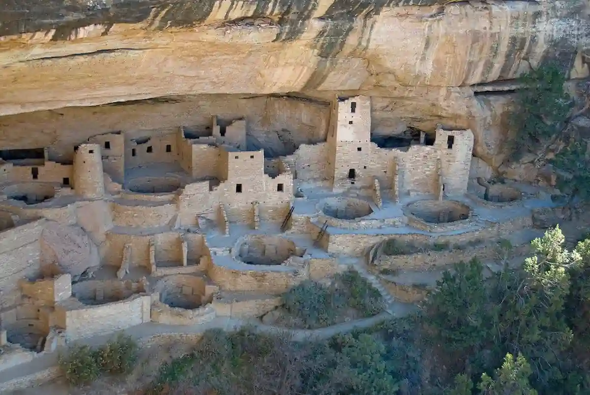 A cliff dwelling with multiple stone structures and circular kivas built into the side of a sandstone cliff, surrounded by greenery at the base. This ancient Puebloan village is a well-preserved archaeological site with terraced levels and rectangular towers.
