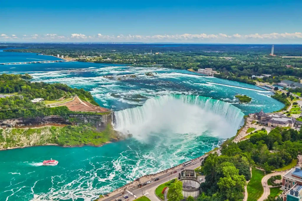 Aerial view of Niagara Falls. The massive, horseshoe-shaped waterfall is surrounded by lush greenery, with mist rising from the cascading waters. A boat with tourists approaches the base of Niagara Falls, while buildings and roads are visible in the background.