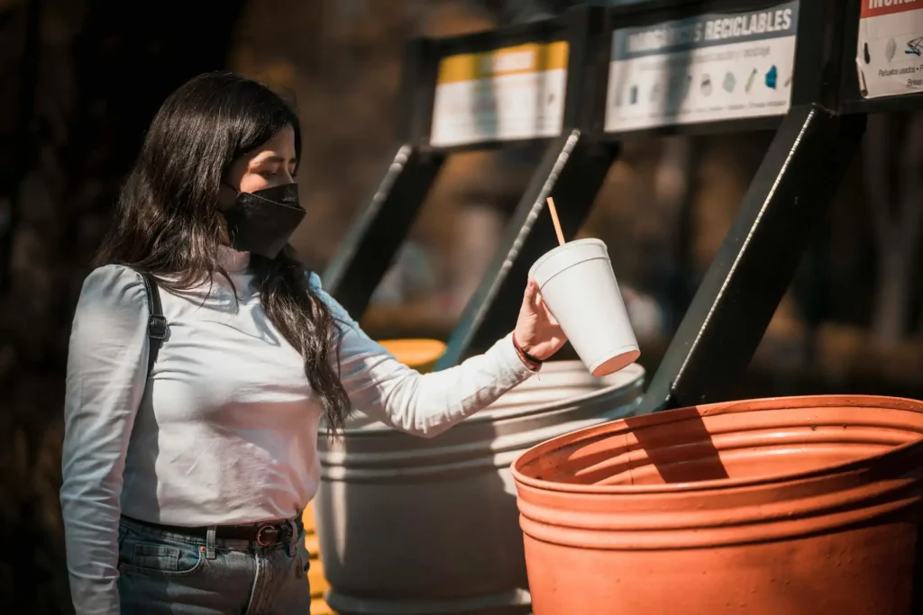 A woman wearing a mask stands in front of a recycling station, holding a white disposable cup with a straw over an orange recycling bin. She is wearing a white long-sleeve shirt and jeans, practicing sustainable travel. The recycling station has labeled containers in the background.