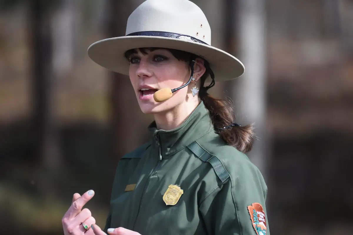 A park ranger wearing a wide-brimmed hat and uniform speaks, pointing with her left hand. She is equipped with a headset microphone for amplification. The background is blurred, with trees indicated in the setting.
