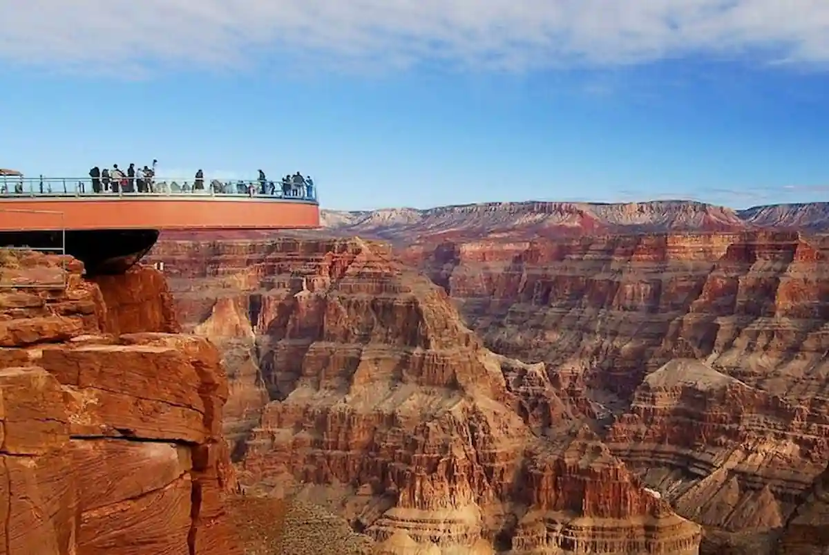 Visitors stand on a glass skywalk extending over the edge of the Grand Canyon, offering panoramic views of the vast, layered rock formations under a blue sky.
 