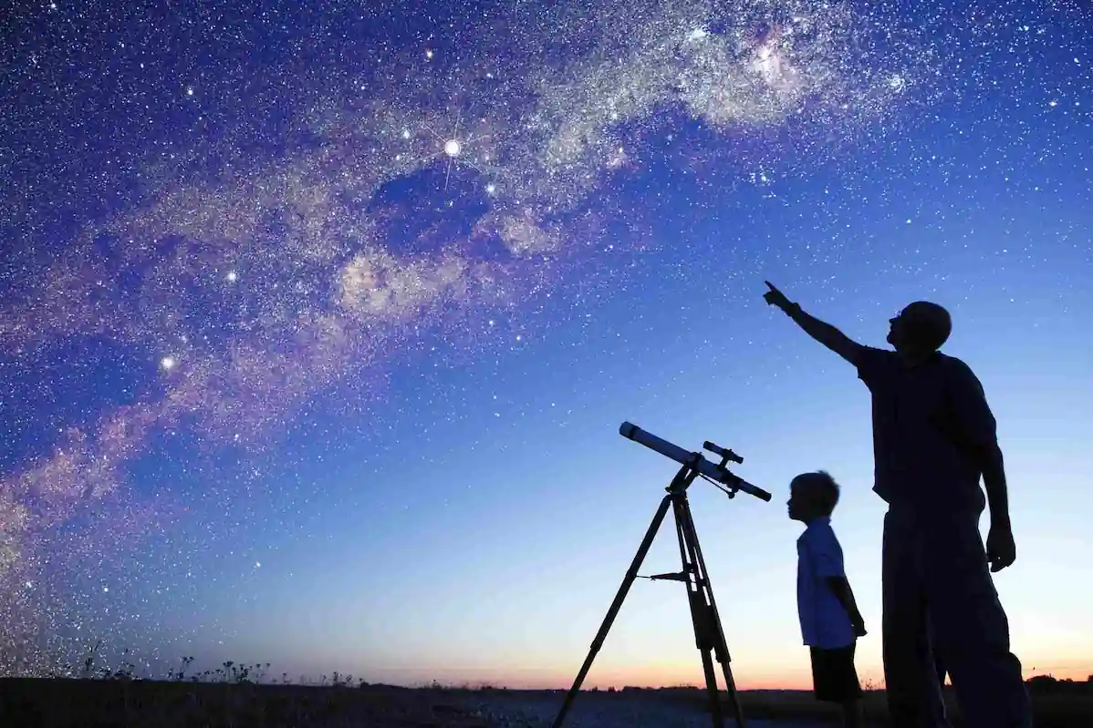 A silhouette of an adult and a child stands next to a telescope under a clear night sky filled with stars and the Milky Way galaxy. The adult points towards the sky while the child looks through the telescope.
