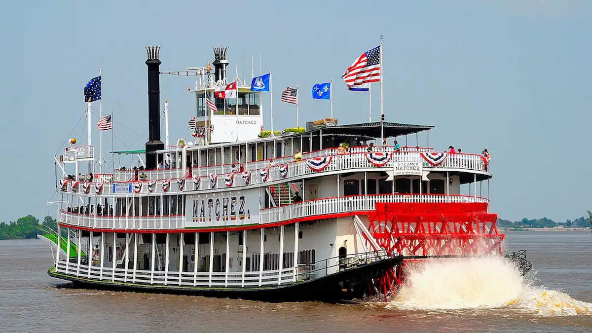 A large, white paddlewheel steamboat with red accents and flags sails on a river. The boat, named "Natchez," has multiple decks adorned with red, white, and blue bunting. Water is being churned by the large red paddlewheel at the back. Trees are visible in the distance.
