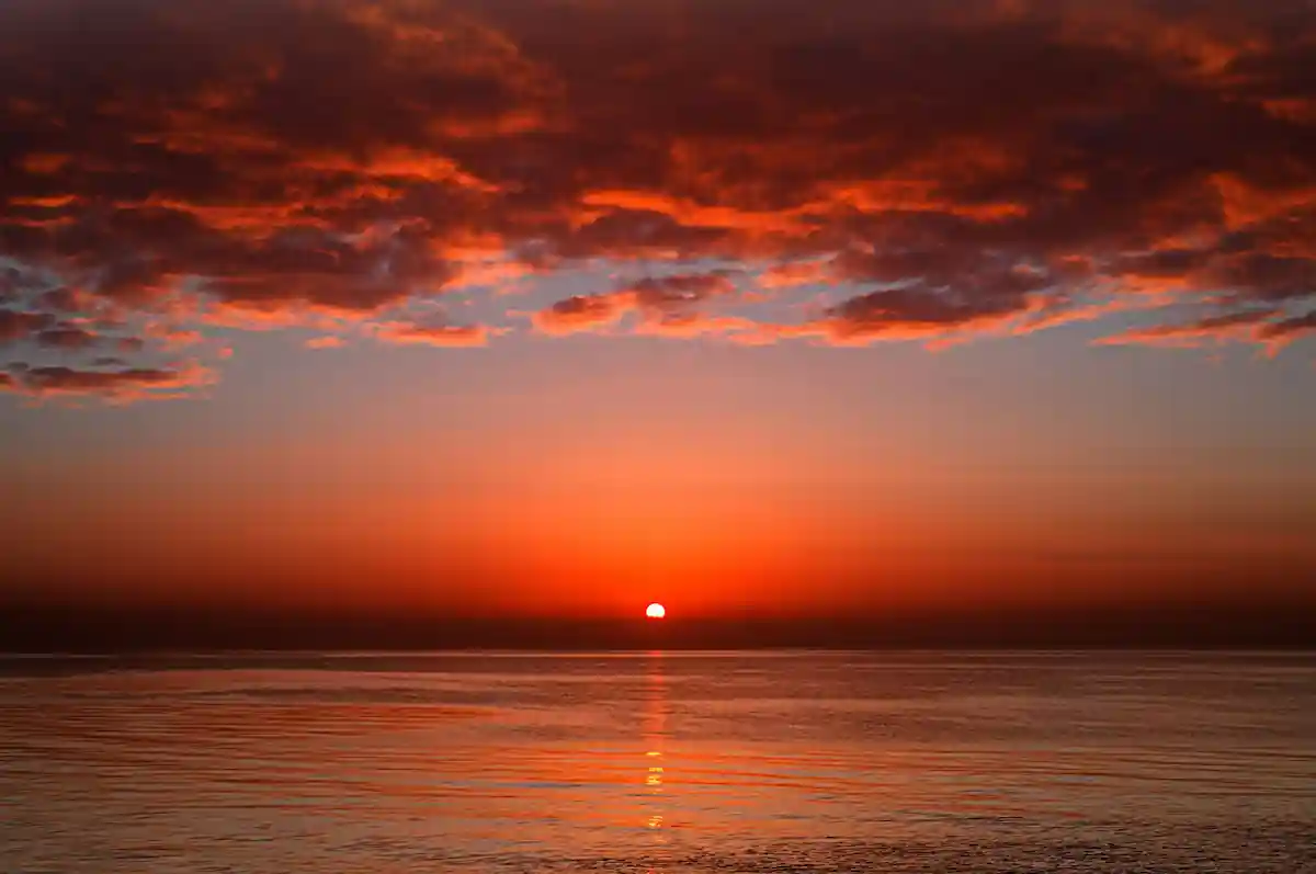 A vivid sunset over a calm ocean, with the sun just above the horizon partially covered by dramatic dark clouds tinged with red and orange hues. A streak of sunlight reflects off the water, creating a serene and captivating scene.
