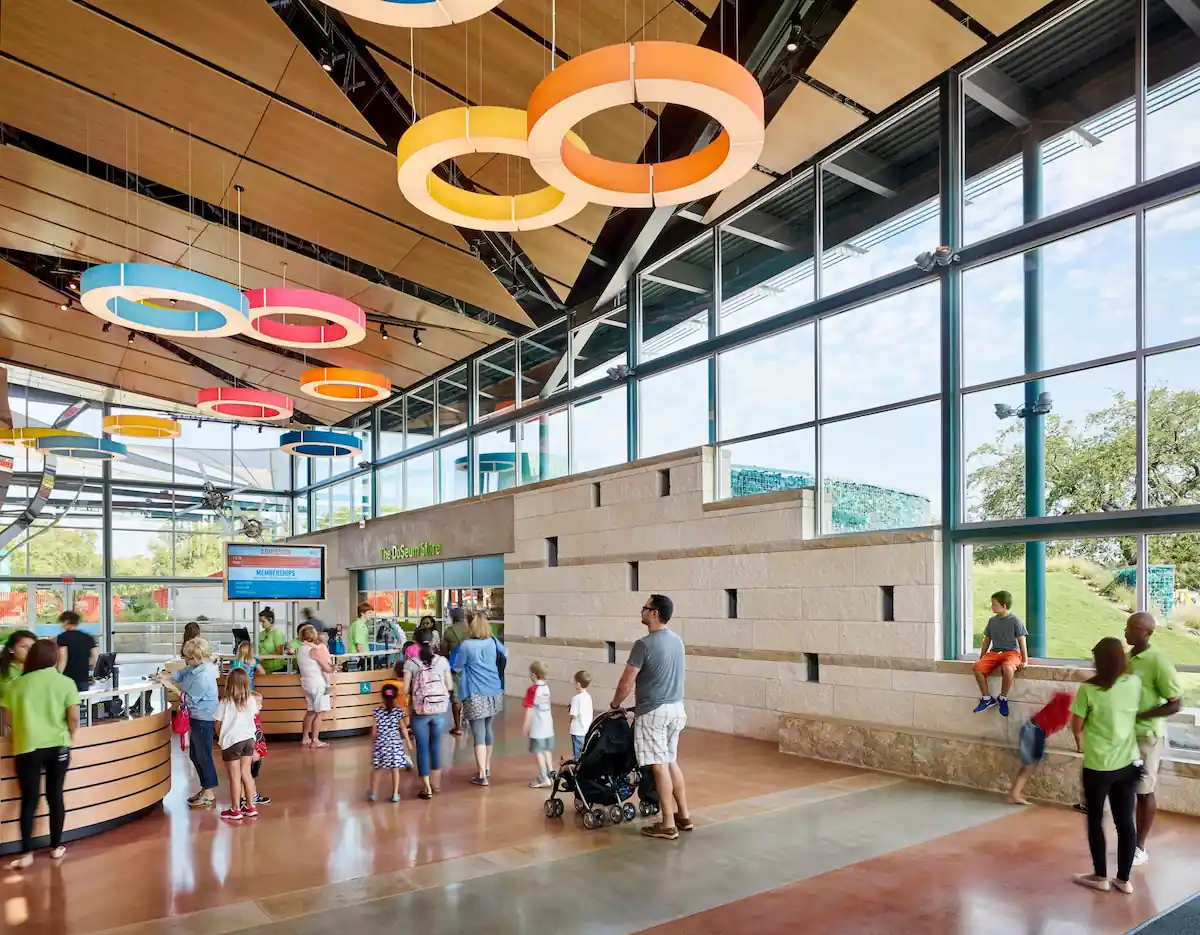 A spacious, modern indoor area with large glass windows and circular light fixtures hanging from the ceiling. Numerous people including families with children are exploring the space. Some children are sitting or playing near a stone wall on the right side.
