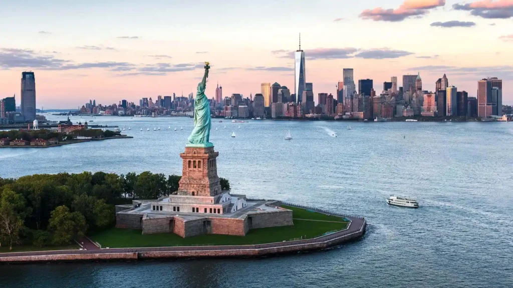 An aerial view captures the Statue of Liberty on Liberty Island, backdropped by the New York City skyline during sunset. Several boats sail on the water, and One World Trade Center stands prominently among other buildings.