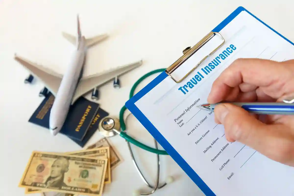 A hand filling out a travel insurance form on a clipboard. Nearby are a model airplane, two passports, scattered US dollar bills, a stethoscope, and a pen. The scene suggests preparing for a trip with consideration for travel and health insurance.
