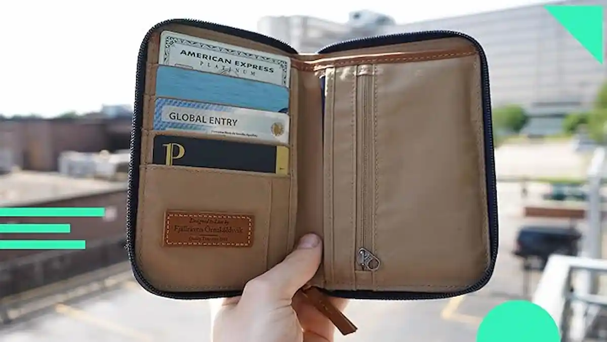 A hand holding an open beige travel wallet in an outdoor setting. The wallet contains various cards such as an American Express card, a Global Entry card, and some other cards organized in slots. The wallet has a zipper and a small brown patch with text.
