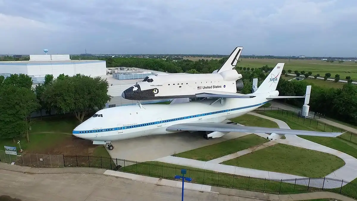 Aerial view of a display at NASA Johnson Space Center, featuring a large airplane with a space shuttle mounted on top. The structure is set against a backdrop of buildings and greenery under a cloudy sky.
