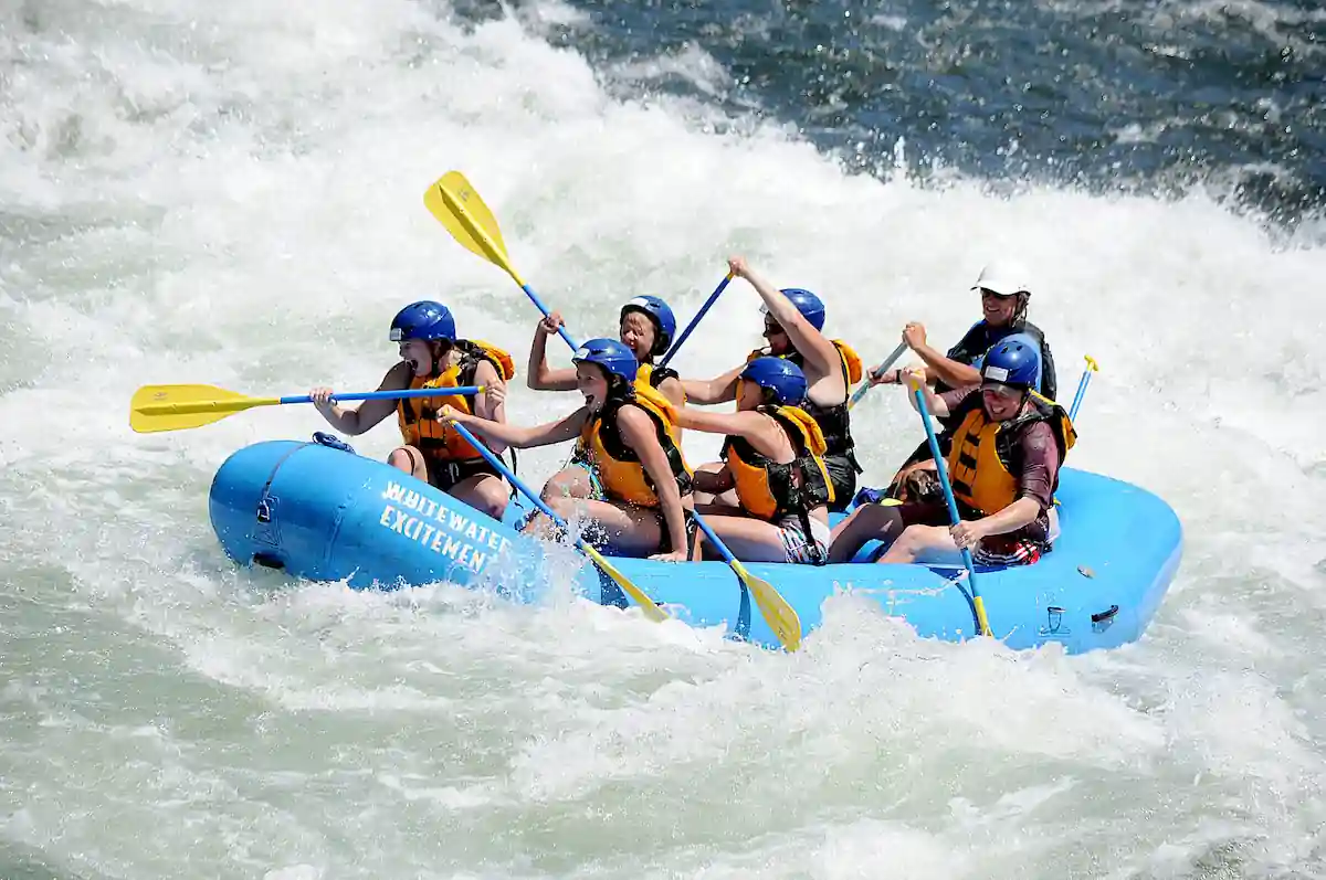 A group of eight people wearing helmets and life vests navigate rough waters in an inflatable blue raft while whitewater rafting. Some paddle vigorously while others hold on, and the water splashes around them, creating an exciting and dynamic scene.
