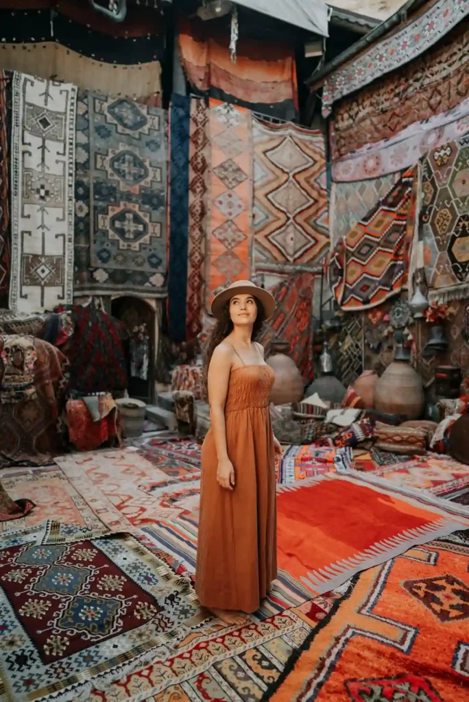 A woman in a long, orange dress and hat stands in a room filled with colorful, patterned rugs and textiles hanging on the walls and covering the floor. She looks up with a thoughtful expression, perhaps contemplating her recent culture travel. The decor features intricate designs and various earthy tones.