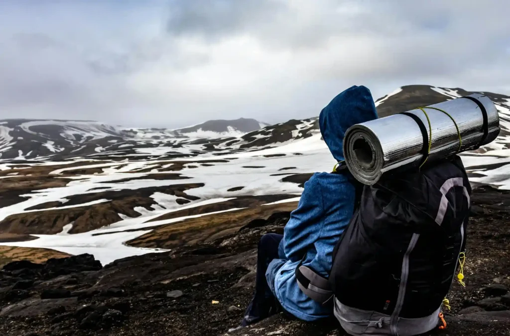 A person wearing a blue jacket and hood sits on a rocky terrain, gazing at a snow-dusted mountainous landscape. Embarking on their solo travel adventure, they have a large backpack with a rolled-up sleeping mat attached to it. The sky is overcast and the scenery looks cold and rugged.