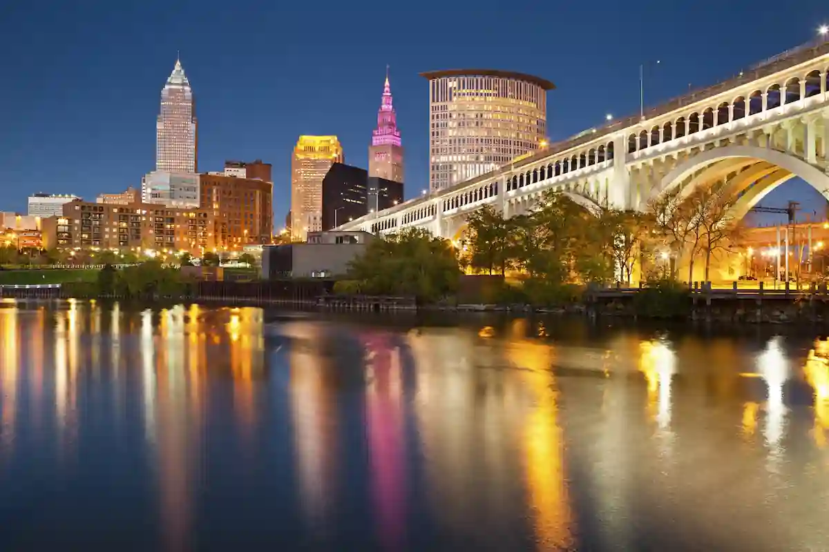 A breathtaking night view of Cleveland, Ohio, showcasing its iconic skyline illuminated against a dark blue sky. The city's skyscrapers, including the towering Key Tower, are reflected in the calm waters of the Cuyahoga River. The iconic Cleveland Memorial Shoreway Bridge, with its arching steel structure, adds to the dramatic scene. This photo captures the beauty and grandeur of Cleveland, making it a must-visit destination for photography enthusiasts seeking stunning urban landscapes. Cleveland skyline, Ohio, cityscape, photography, night photography, urban landscape, Key Tower, Cleveland Memorial Shoreway Bridge, Cuyahoga River, beautiful places in Ohio to take pictures, Ohio travel destinations, Cleveland attractions, night photography tips, urban photography, must-visit places in Ohio