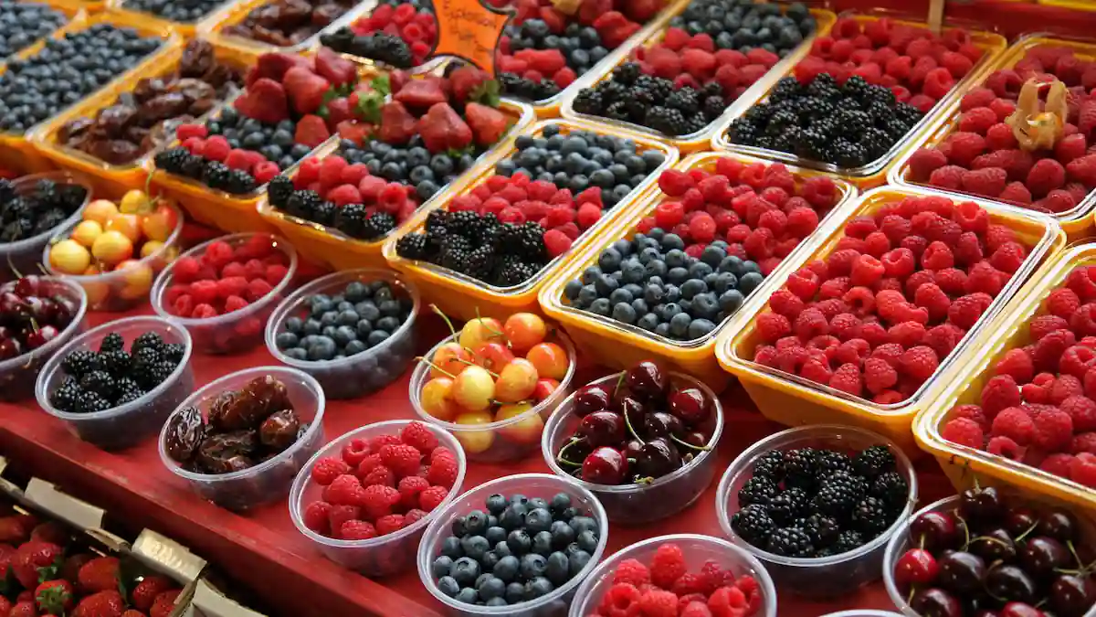 A colorful display of various fruits at a market, featuring containers filled with strawberries, blackberries, blueberries, raspberries, cherries, and dates, all arranged neatly in rows. A small yellow sign with handwritten text is visible in the background.
