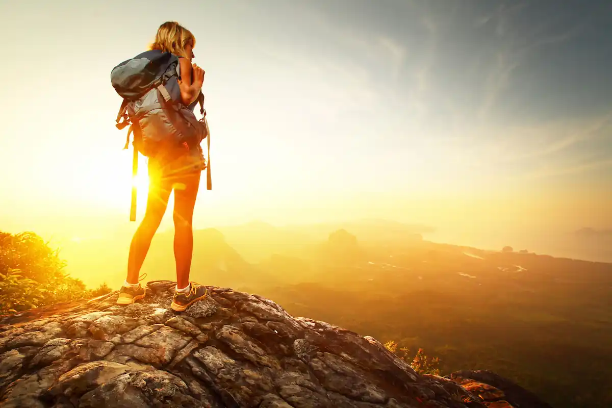 A hiker stands on a rocky cliff, overlooking a vast landscape. They are wearing a backpack and using binoculars. The sun is setting, casting a golden glow across the scenery, with mountains and valleys visible in the distance.
 