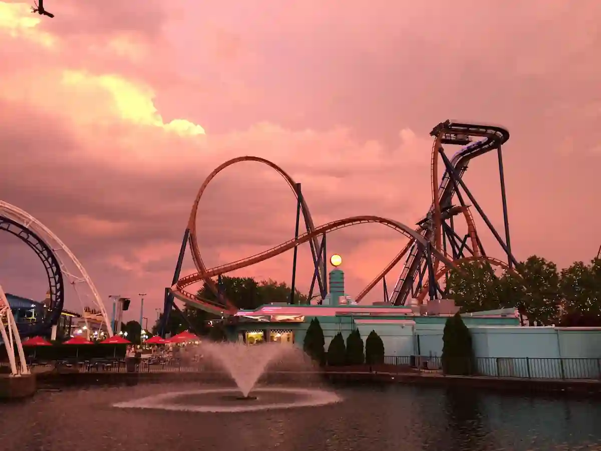 A thrilling roller coaster at Cedar Point amusement park stands tall against a dramatic sunset sky. The rollercoaster's tracks twist and turn, leading to a heart-pounding experience. A fountain sprays water in the foreground, adding a touch of serenity to the scene. The image evokes a sense of excitement and adventure.