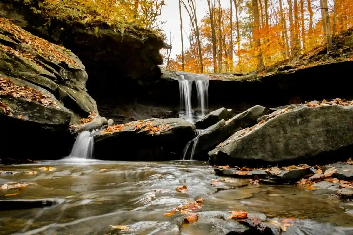 A scenic waterfall cascades down a rocky cliff, surrounded by vibrant autumn foliage. Sunlight filters through the leaves, casting dappled shadows on the water and rocks below. The scene evokes a sense of tranquility and natural beauty.