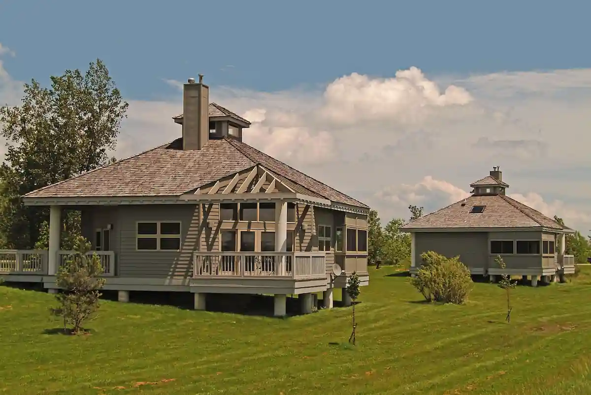 Two cozy cabins with shingled roofs and large windows are nestled on a grassy hill. The cabins feature spacious decks overlooking a scenic landscape. The surrounding area is lush and green, with mature trees and a clear blue sky with fluffy clouds.