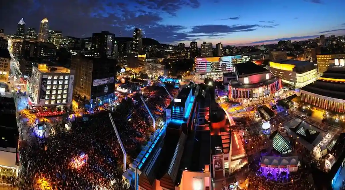 A vibrant night scene of a city festival with a large crowd gathered. Colorful lights illuminate the streets and buildings, creating an energetic atmosphere. The city skyline is visible in the background against a deep blue night sky.
