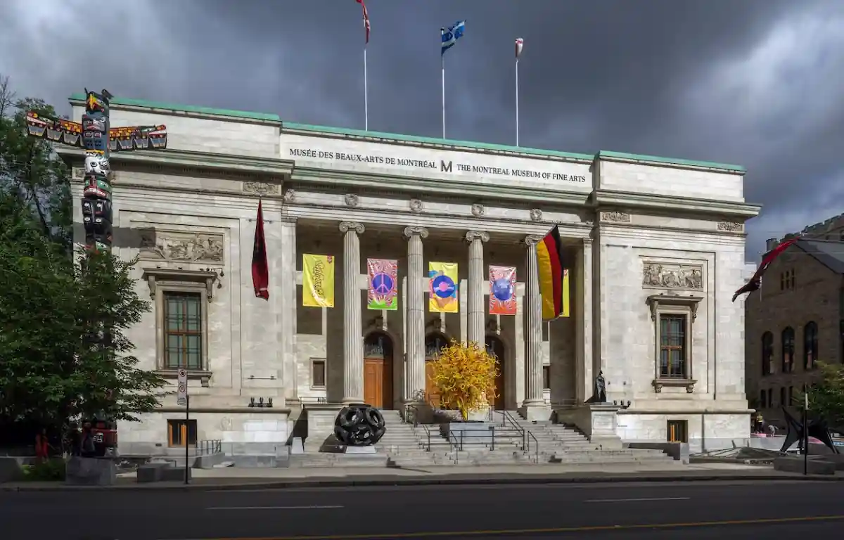 Front view of the Montreal Museum of Fine Arts under a dark cloudy sky. The building has grand columns and banners hanging between them. A decorative yellow sculpture sits on the steps, and a totem pole stands to the left of the entrance.
