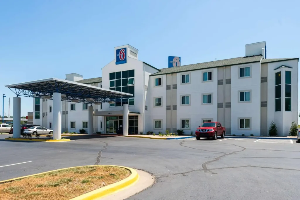 A white, two-story Motel 6 building with a blue awning over the entrance. The building has multiple windows and a large, rectangular sign featuring the Motel 6 logo and a cartoon dog. Cars are parked in the motel's parking lot. The motel is located in a suburban area with visible road signs and power lines.