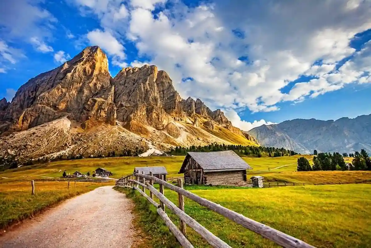 A scenic mountain landscape featuring a dirt path lined with a wooden fence leading to rustic cabins. The backdrop showcases rugged mountains under a partly cloudy sky, with lush green fields in the foreground.
