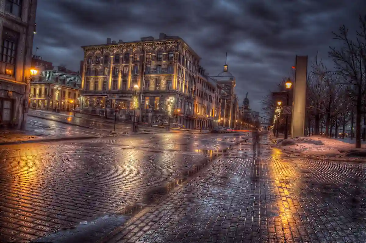 A rainy, night-time cityscape with wet cobblestone streets reflecting warm yellow lights from surrounding buildings. An elegant, historic building with lit windows stands prominently on the left, while dark, dramatic clouds loom overhead.
