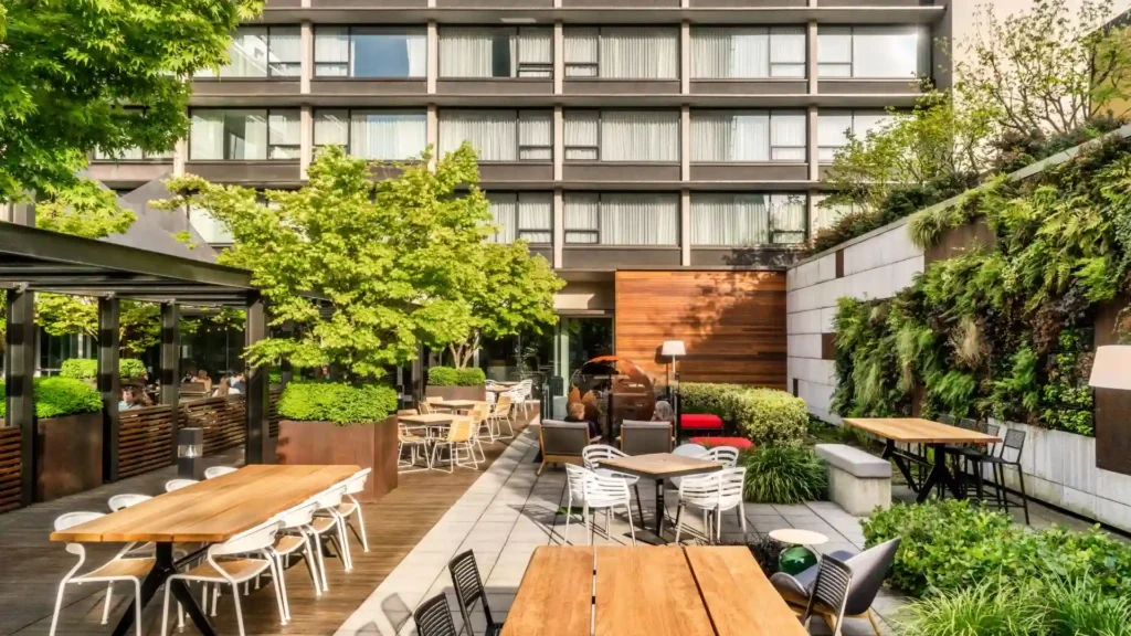  A photo of the Hotel Zags Portland's outdoor patio. The patio features a large, rectangular wooden table with chairs, a fire pit, and lush greenery, including trees, shrubs, and a vertical garden. There is also a covered seating area with a glass roof. The hotel building is visible in the background.