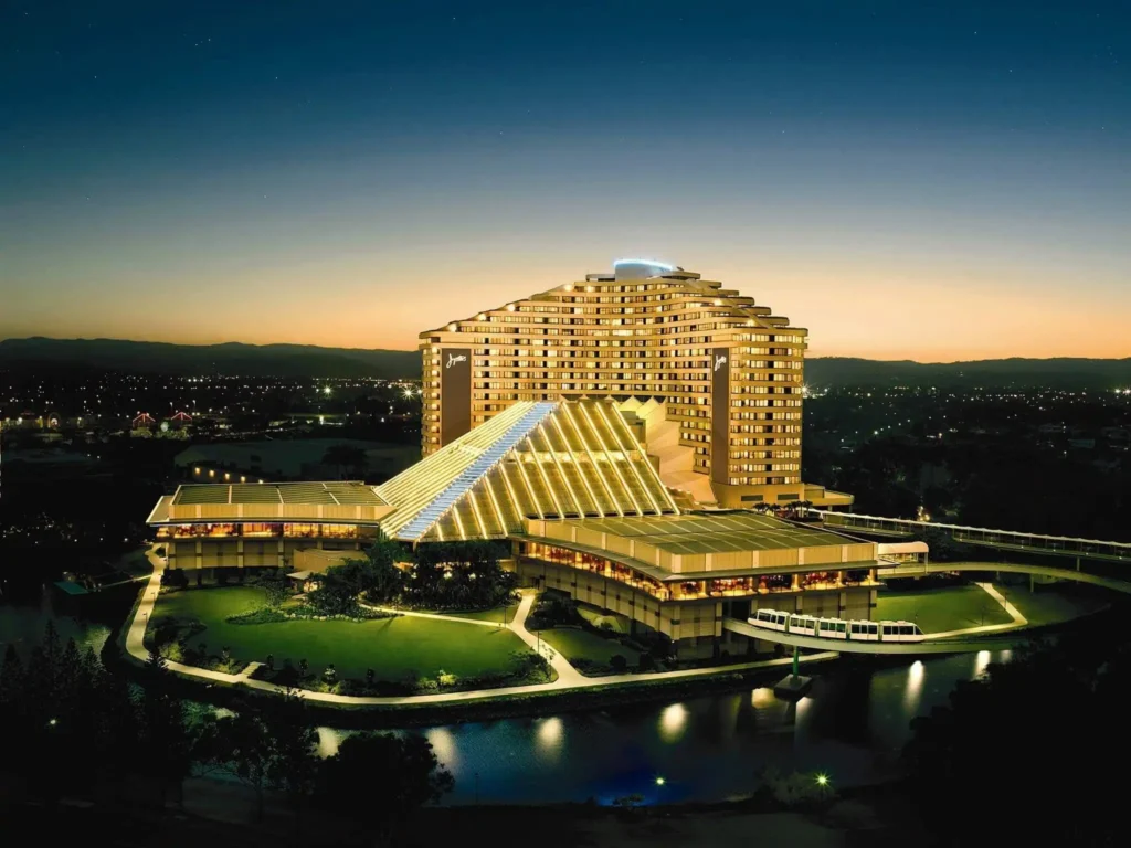 A large, illuminated hotel complex with a distinctive pyramid-shaped tower. The hotel is surrounded by landscaped grounds and is located near a body of water. A monorail system runs along the property. The image likely depicts the Jupiter's Casino and Hotel in Gold Coast, Australia.