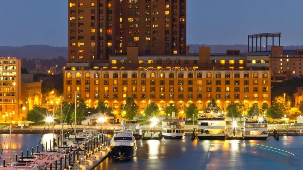 A photo of the Royal Sonesta Harbor Court Baltimore at night. The hotel is a large, illuminated building with a distinctive facade and is located on the waterfront. There are boats docked in the marina, and the city skyline is visible in the background.