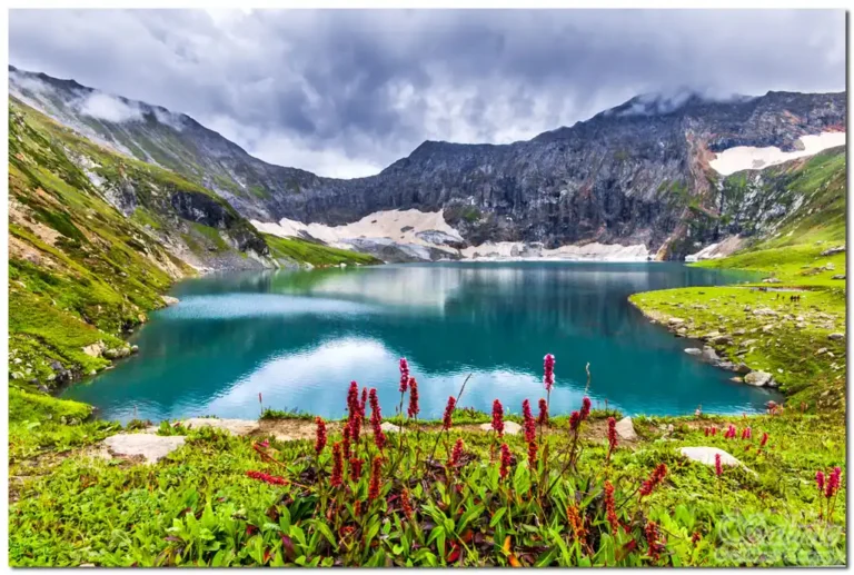 A tranquil mountain lake surrounded by rocky peaks and patches of snow under a cloudy sky. In the foreground, vibrant red wildflowers and green grass frame the serene blue waters. Ratti Gali Laken is one of the Places That Do Not Feel Real Pakistan